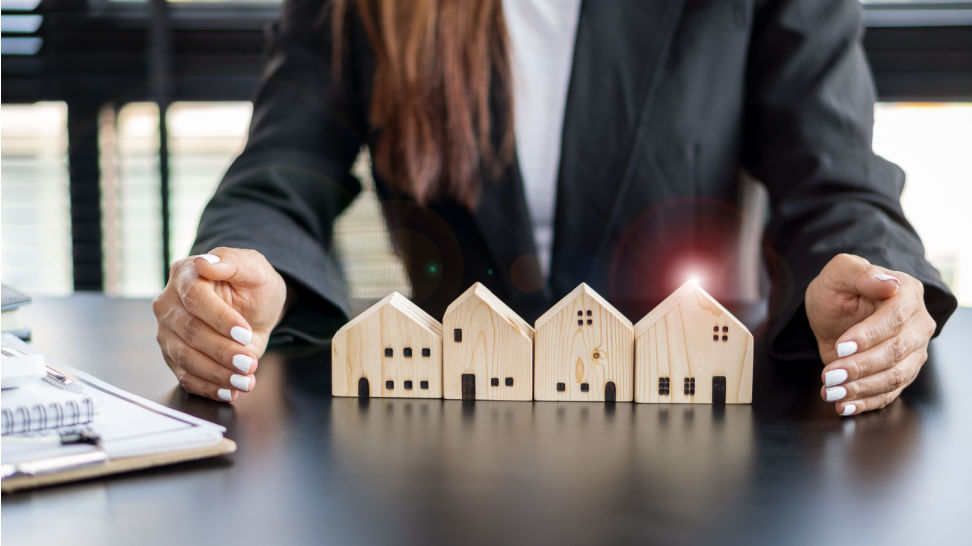 Businesswoman in a dark suit cupping her hands protectively around four small wooden model houses on a desk